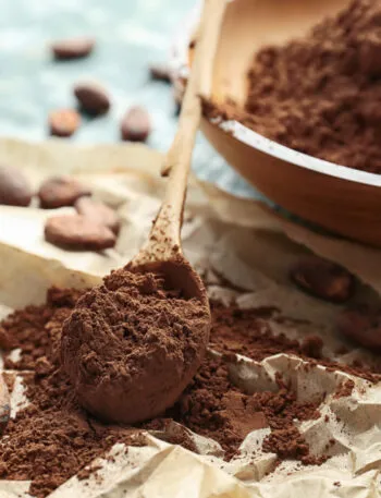 Cacao amaro in polvere su un cucchiaio di legno, con ciotola e fave di cacao su un tavolo azzurro.