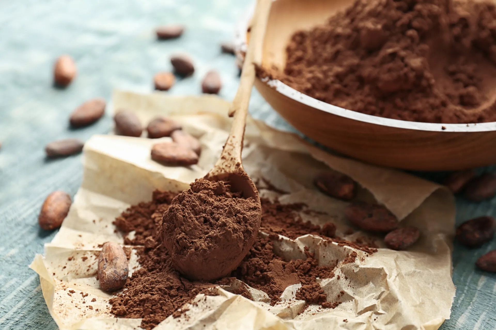 Cacao amaro in polvere su un cucchiaio di legno, con ciotola e fave di cacao su un tavolo azzurro.