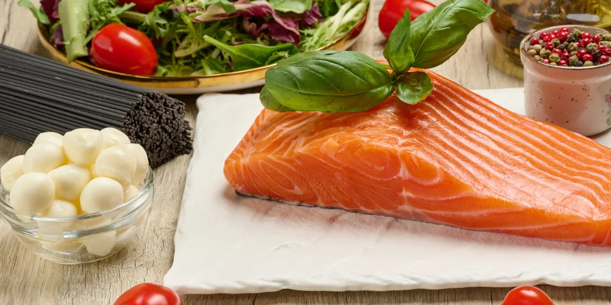 Filetto di salmone fresco con basilico, pomodorini, insalata, mozzarella e pasta integrale su tavolo in legno.
