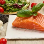 Filetto di salmone fresco con basilico, pomodorini, insalata, mozzarella e pasta integrale su tavolo in legno.