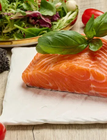 Filetto di salmone fresco con basilico, pomodorini, insalata, mozzarella e pasta integrale su tavolo in legno.