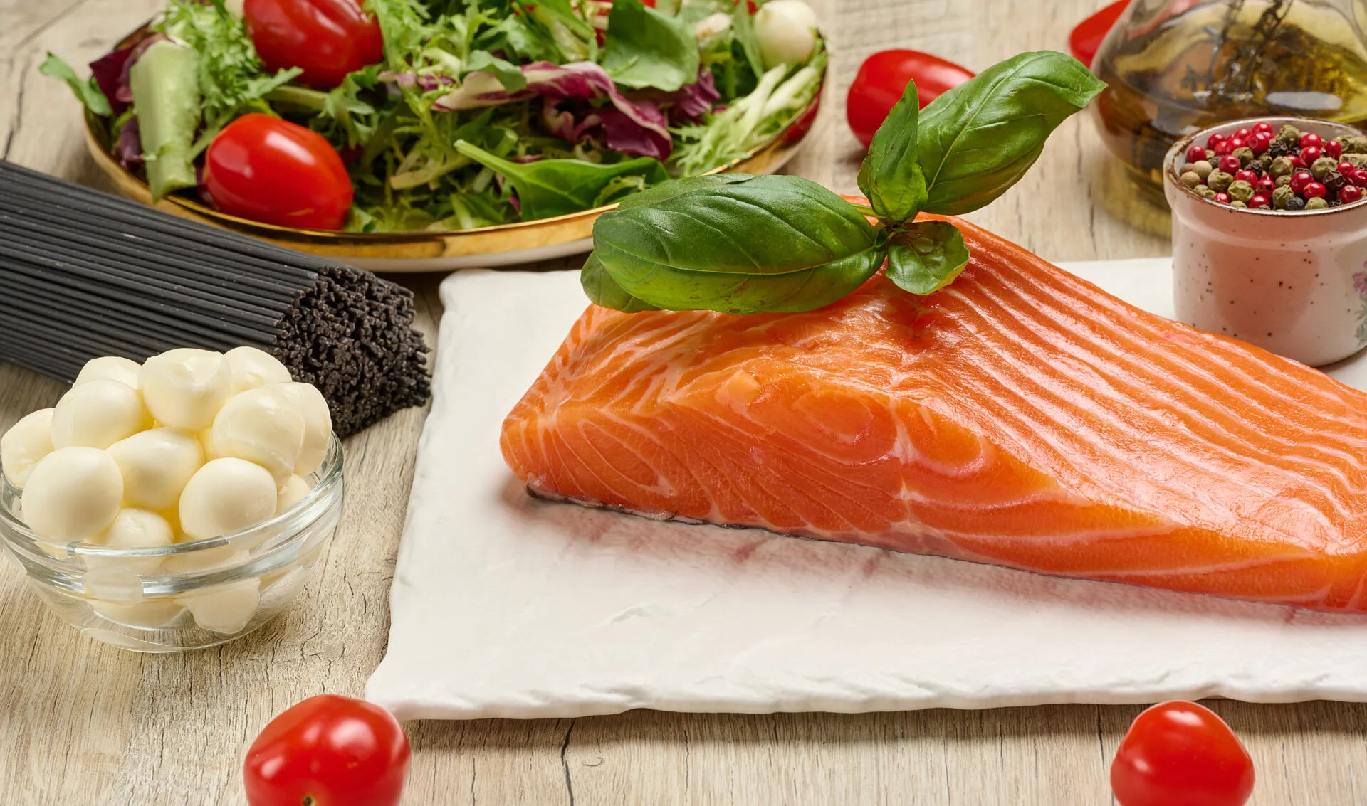 Filetto di salmone fresco con basilico, pomodorini, insalata, mozzarella e pasta integrale su tavolo in legno.