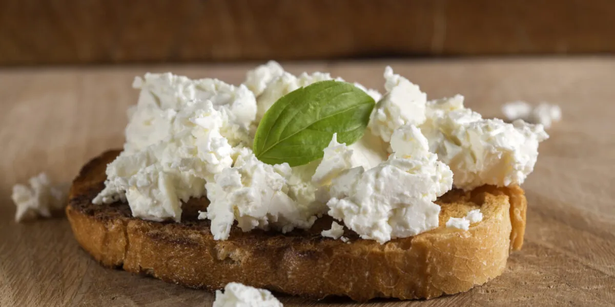 Fetta di pane tostato con ricotta fresca e foglia di basilico su tagliere di legno.