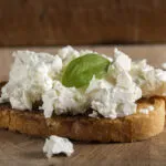 Fetta di pane tostato con ricotta fresca e foglia di basilico su tagliere di legno.