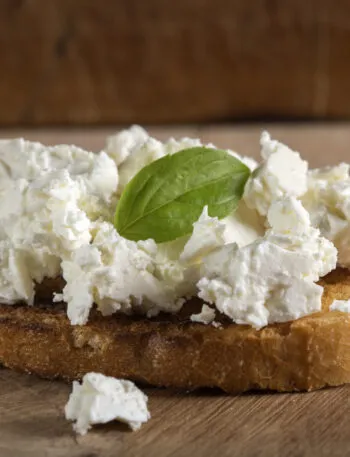 Fetta di pane tostato con ricotta fresca e foglia di basilico su tagliere di legno.