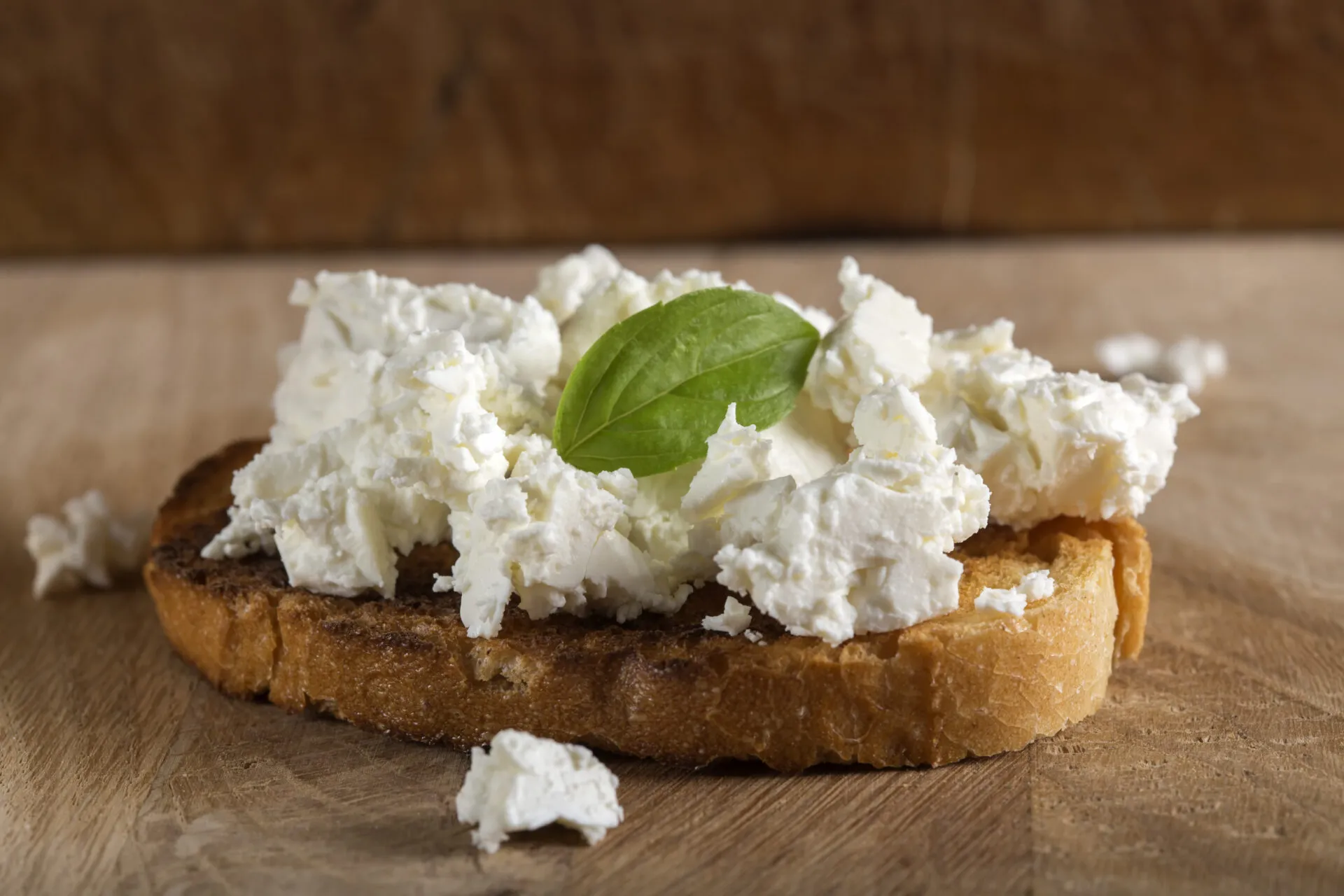 Fetta di pane tostato con ricotta fresca e foglia di basilico su tagliere di legno.