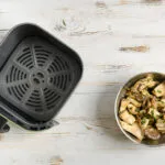 Friggitrice ad aria aperta con ciotola di funghi conditi su un tavolo in legno bianco.