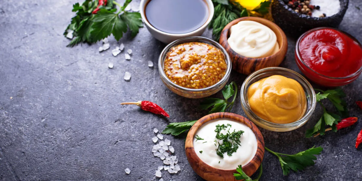 Salse fit fatte in casa in ciotole con ketchup, maionese, senape, yogurt, spezie ed erbe aromatiche su fondo scuro.