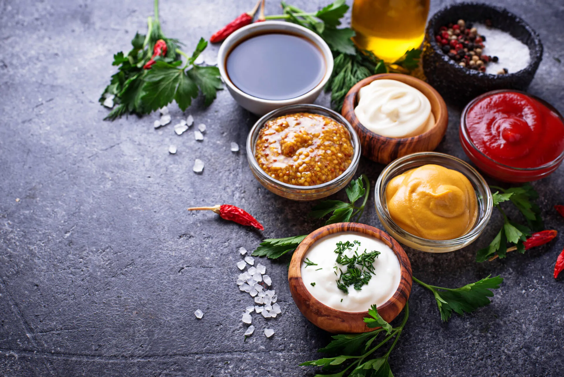 Salse fit fatte in casa in ciotole con ketchup, maionese, senape, yogurt, spezie ed erbe aromatiche su fondo scuro.