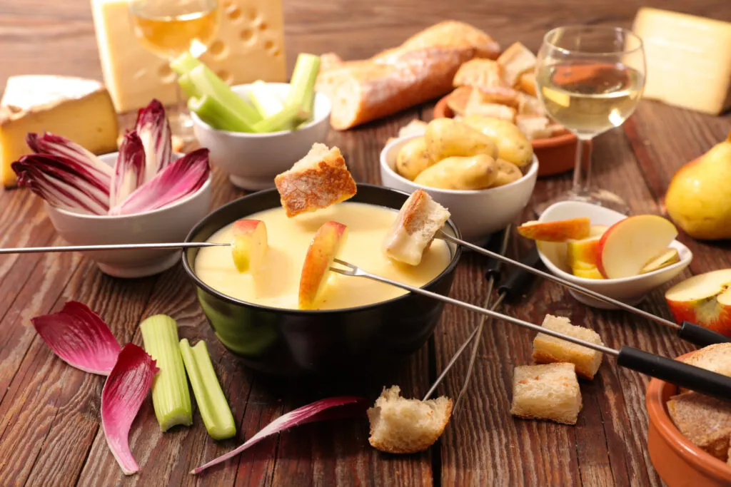 Set da fonduta con formaggio fuso, pane, mele, patate, verdure e calici di vino su un tavolo di legno.