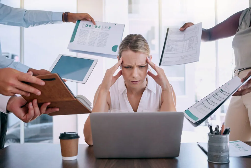 Donna stressata al lavoro davanti al computer mentre colleghi le mostrano documenti, situazione che può favorire la comparsa della fame nervosa.