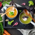 Due vellutate fit, una arancione e una verde, servite con yogurt ed erbe aromatiche, circondate da carote, broccoli, cavolfiore e spezie.