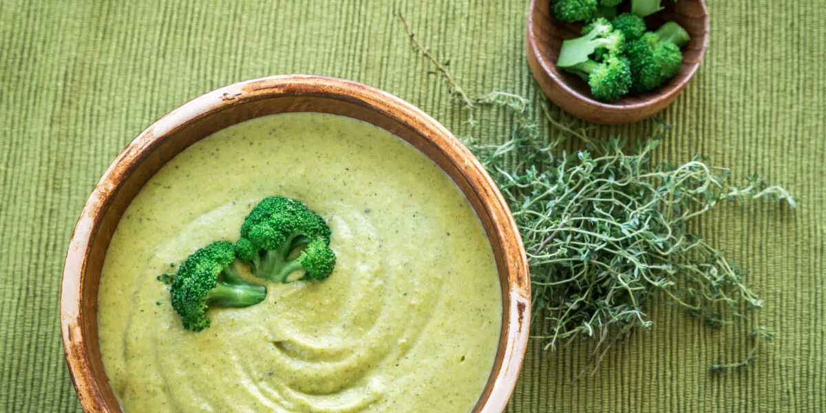 Crema di broccoli vellutata servita in una ciotola, decorata con cimette verdi e accompagnata da broccoli e erbe aromatiche.