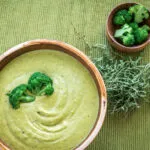 Crema di broccoli vellutata servita in una ciotola, decorata con cimette verdi e accompagnata da broccoli e erbe aromatiche.