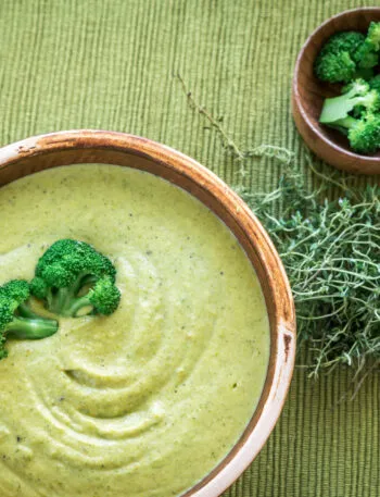 Crema di broccoli vellutata servita in una ciotola, decorata con cimette verdi e accompagnata da broccoli e erbe aromatiche.