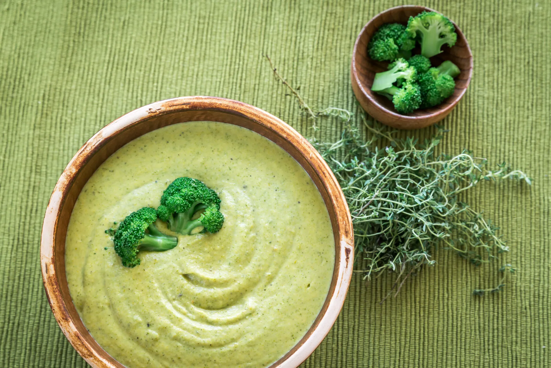 Crema di broccoli vellutata servita in una ciotola, decorata con cimette verdi e accompagnata da broccoli e erbe aromatiche.