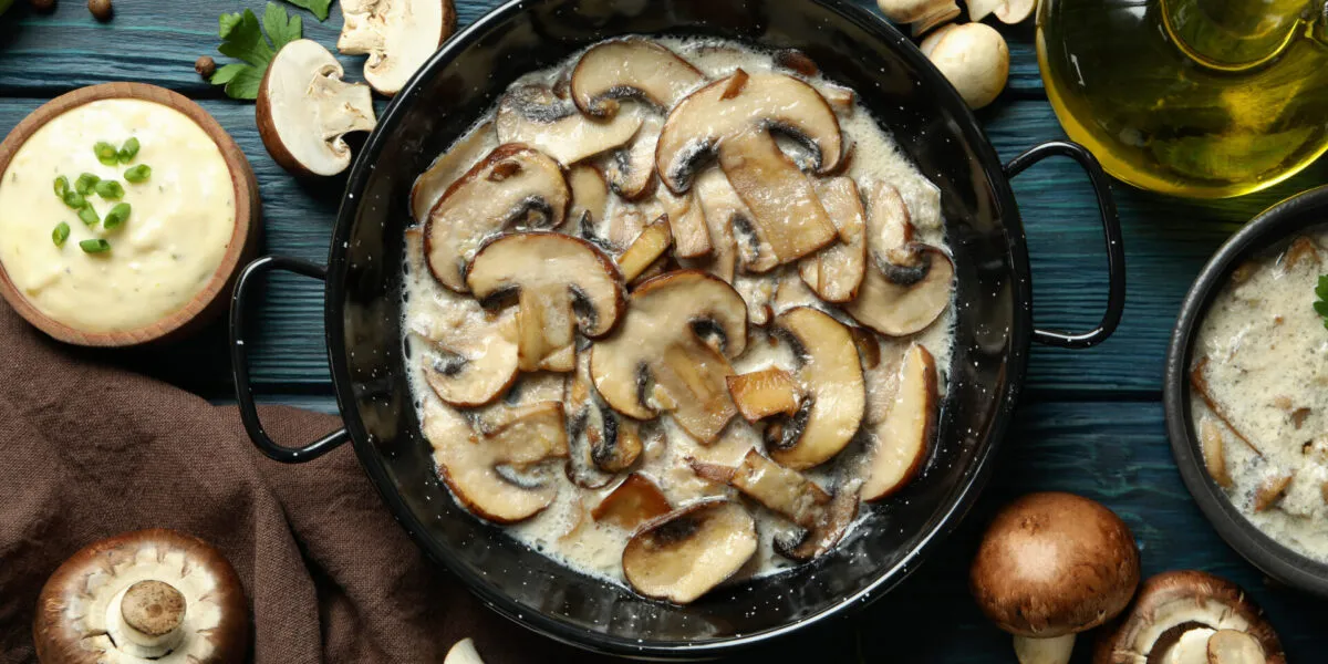 Funghi champignon affettati in padella con salsa cremosa, prezzemolo, olio e funghi freschi su tavolo di legno blu.