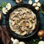 Funghi champignon affettati in padella con salsa cremosa, prezzemolo, olio e funghi freschi su tavolo di legno blu.