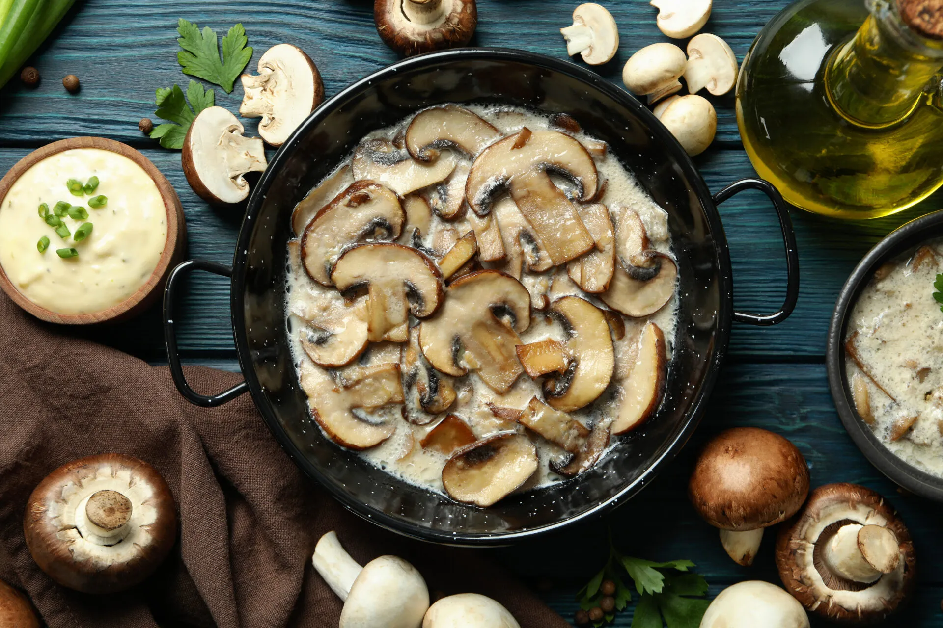 Funghi champignon affettati in padella con salsa cremosa, prezzemolo, olio e funghi freschi su tavolo di legno blu.