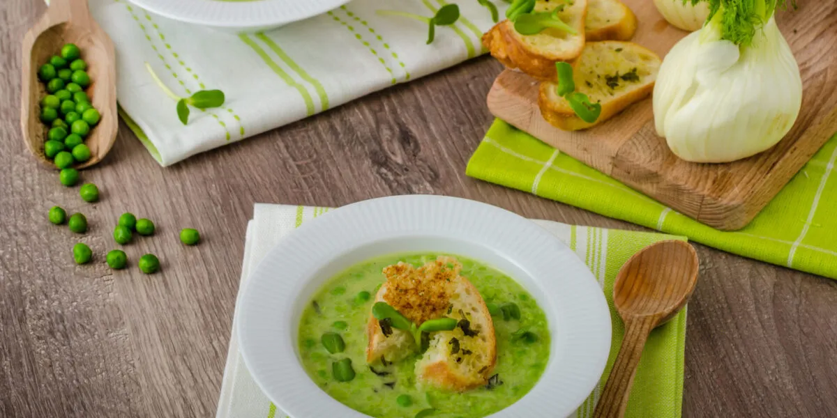 Vellutata di piselli con crostini, piselli freschi e finocchi su un tavolo in legno.