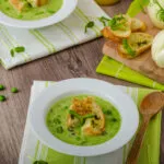 Vellutata di piselli con crostini, piselli freschi e finocchi su un tavolo in legno.
