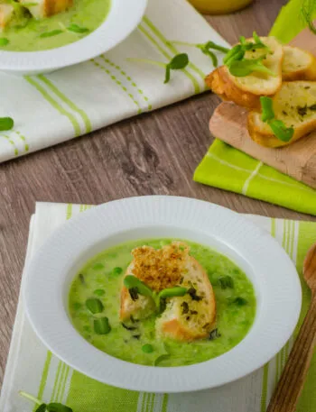 Vellutata di piselli con crostini, piselli freschi e finocchi su un tavolo in legno.