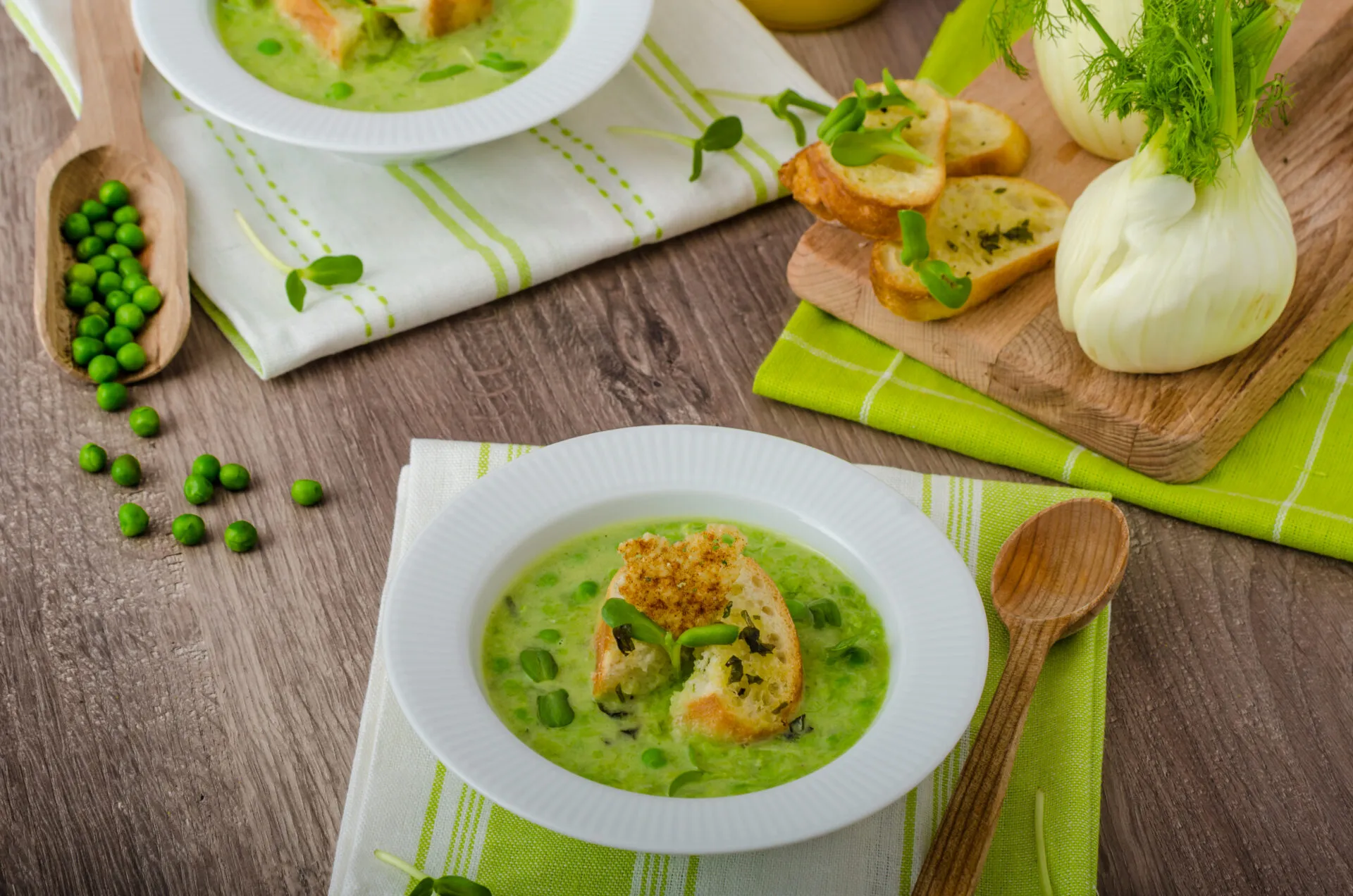 Vellutata di piselli con crostini, piselli freschi e finocchi su un tavolo in legno.