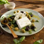 Feta con olive verdi e nere, basilico fresco, olio ed erbe aromatiche su un piatto.