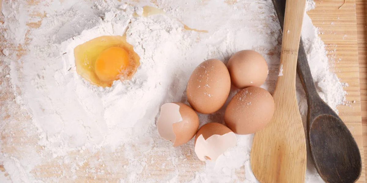 Uova intere e gusci accanto a un uovo rotto nella farina, con cucchiai di legno su un piano da cucina.