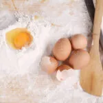 Uova intere e gusci accanto a un uovo rotto nella farina, con cucchiai di legno su un piano da cucina.