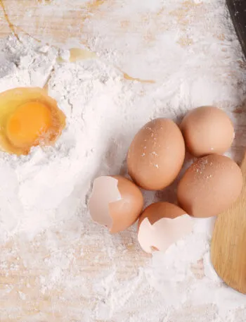 Uova intere e gusci accanto a un uovo rotto nella farina, con cucchiai di legno su un piano da cucina.