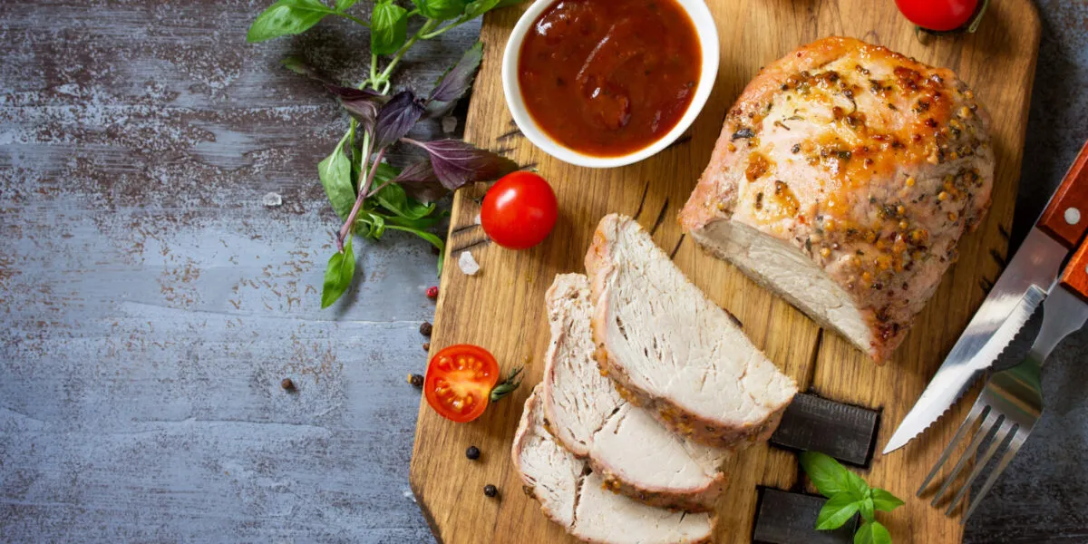 Arrosto di tacchino alle erbe tagliato a fette con salsa, pomodorini e basilico su un tagliere di legno.