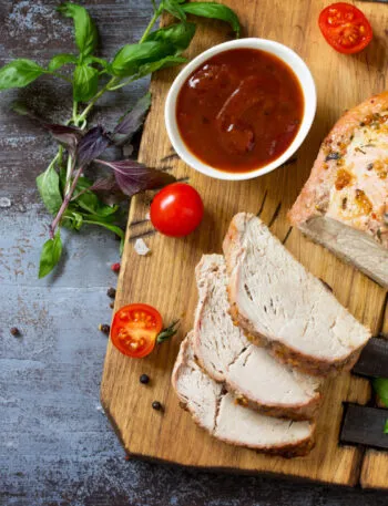 Arrosto di tacchino alle erbe tagliato a fette con salsa, pomodorini e basilico su un tagliere di legno.