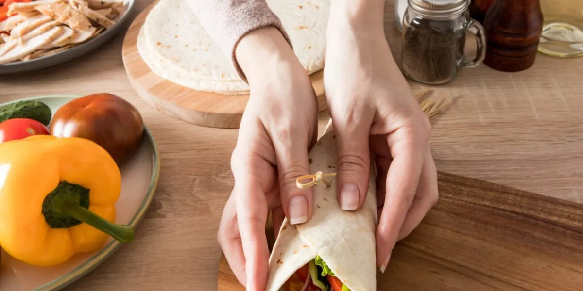 Mani che chiudono un wrap fit ripieno di pollo, lattuga, pomodoro, cetriolo e cipolla rossa.