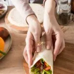Mani che chiudono un wrap fit ripieno di pollo, lattuga, pomodoro, cetriolo e cipolla rossa.