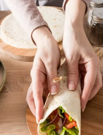 Mani che chiudono un wrap fit ripieno di pollo, lattuga, pomodoro, cetriolo e cipolla rossa.
