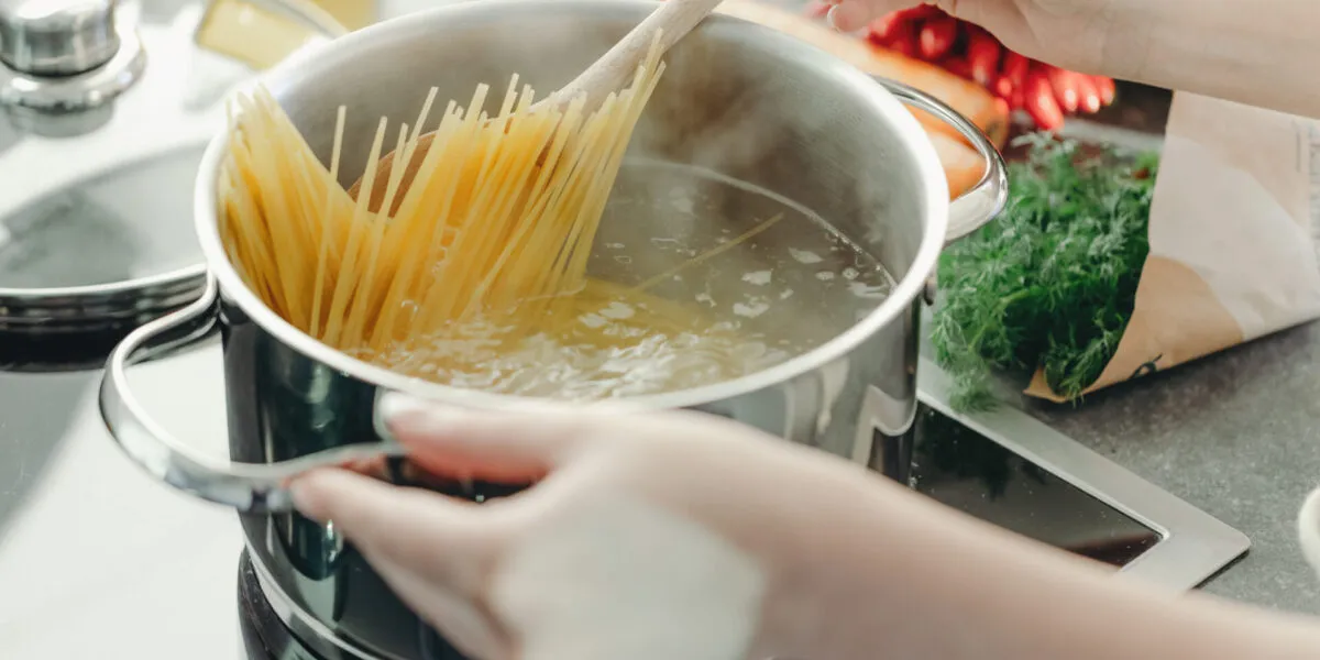 Mani che calano gli spaghetti in una pentola di acqua bollente mescolando con un cucchiaio di legno, su piano cottura a induzione