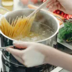 Mani che calano gli spaghetti in una pentola di acqua bollente mescolando con un cucchiaio di legno, su piano cottura a induzione