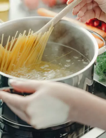 Mani che calano gli spaghetti in una pentola di acqua bollente mescolando con un cucchiaio di legno, su piano cottura a induzione