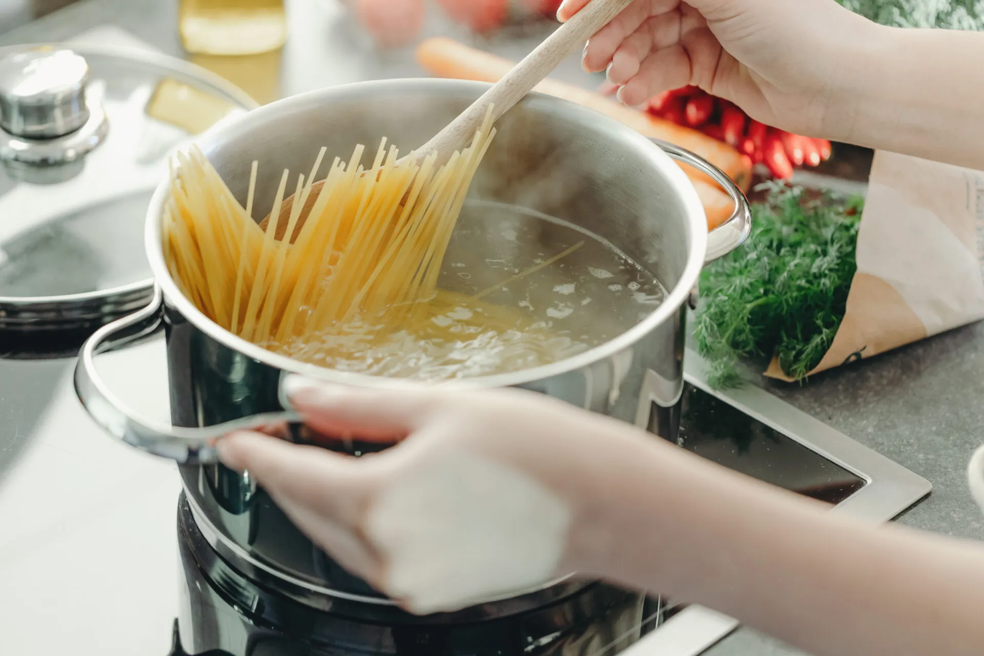 Mani che calano gli spaghetti in una pentola di acqua bollente mescolando con un cucchiaio di legno, su piano cottura a induzione