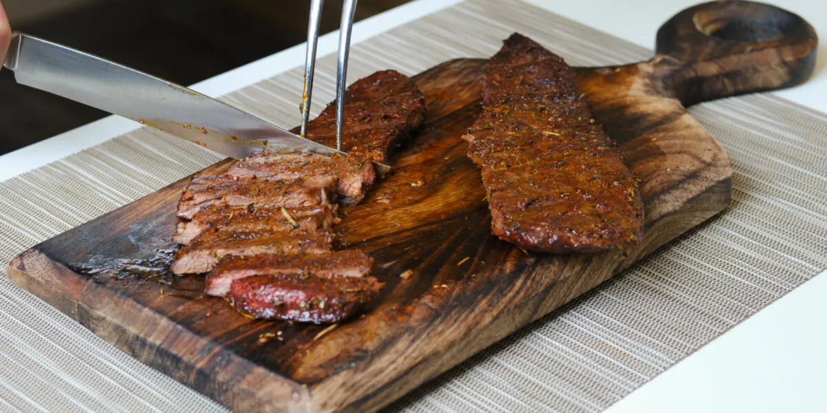 Carne affumicata con crosta di erbe affettata su un tagliere di legno con coltello e forchettone