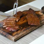 Carne affumicata con crosta di erbe affettata su un tagliere di legno con coltello e forchettone