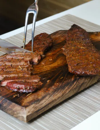 Carne affumicata con crosta di erbe affettata su un tagliere di legno con coltello e forchettone