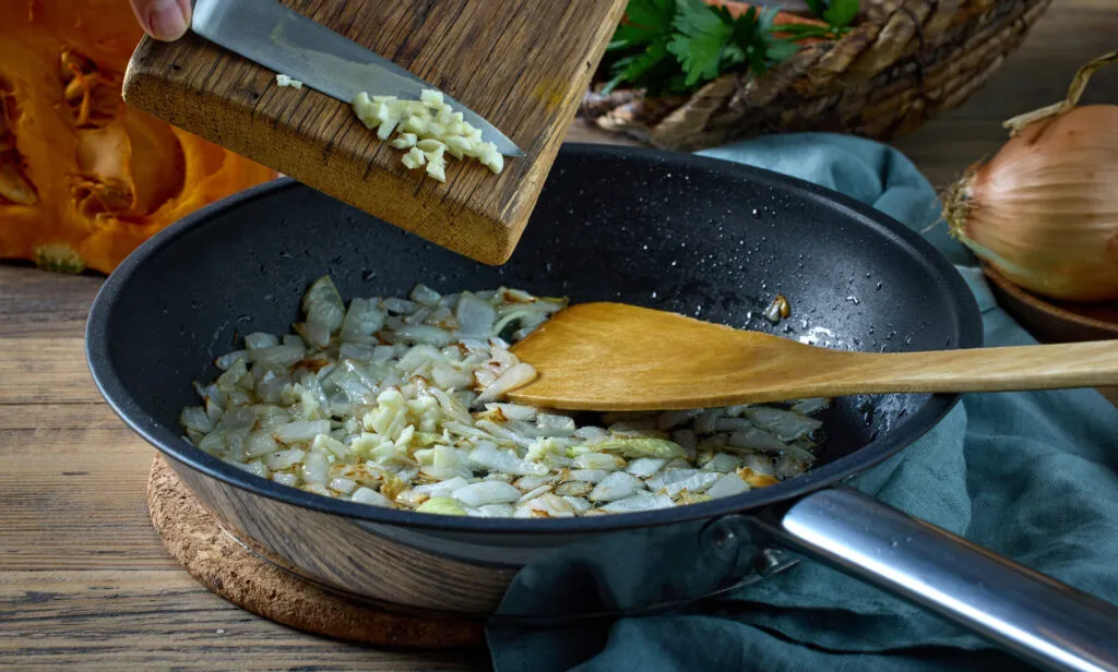 Aglio tritato fatto scivolare dal tagliere in una padella con cipolla che stufa, ancora chiaro e per niente dorato