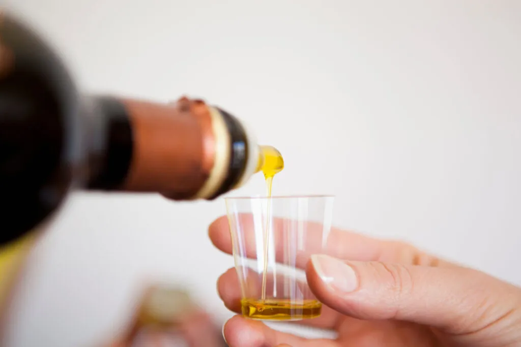 Olio extravergine versato da una bottiglia di vetro scuro in un bicchierino trasparente tenuto in mano per l'assaggio
