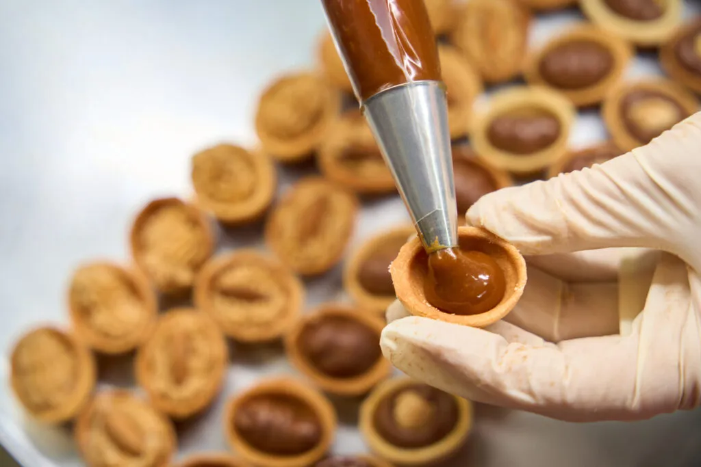 Mano con guanto che farcisce una tartelletta con una sac à poche in un laboratorio di pasticceria, con altre tartellette sul vassoio
