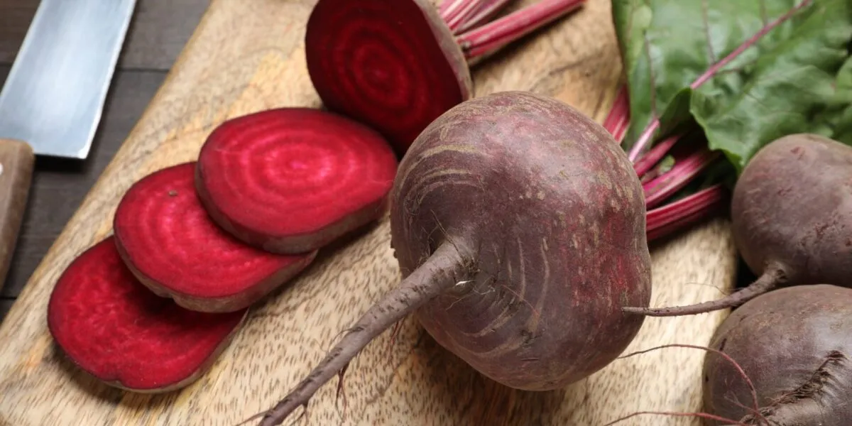 Barbabietole crude su un tagliere di legno, una tagliata a fette che mostrano gli anelli concentrici rosso rubino, con coltello e foglie accanto