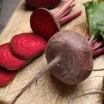 Barbabietole crude su un tagliere di legno, una tagliata a fette che mostrano gli anelli concentrici rosso rubino, con coltello e foglie accanto