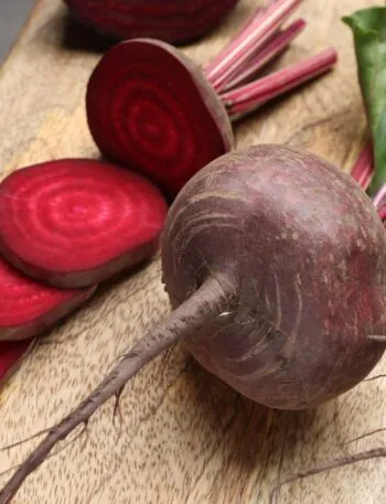 Barbabietole crude su un tagliere di legno, una tagliata a fette che mostrano gli anelli concentrici rosso rubino, con coltello e foglie accanto
