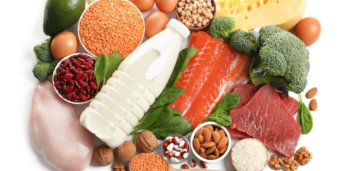 Vista dall’alto di fonti proteiche diverse su fondo bianco: petto di pollo, salmone, uova, legumi, formaggio, frutta secca, riso e broccoli